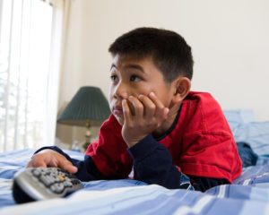 Young asian boy laying down on his stomach on the bed while looking engrossed in the television holding the remote control